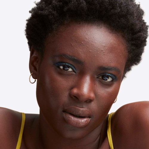 A woman with short curly hair, wearing yellow top straps, with blue eyeshadow and earrings, looks directly at the camera.