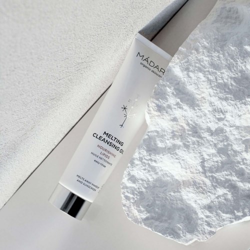 A white tube of Melting Cleansing Oil lies on white textured surface with a pile of cotton around it.