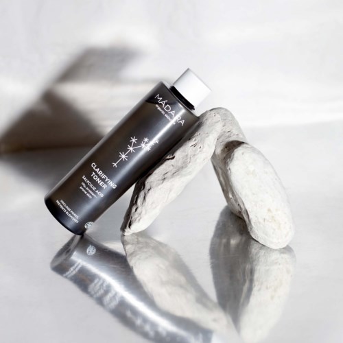 Musta Madara Clarifying Toner -pullo lepää valkoisten kivien päällä, heijastuu peilinä minimalistisessa ympäristössä.