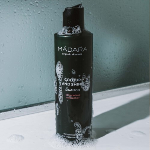 Black bottle of MÁDARA shampoo with white bubbles, placed on a surface with some foam, near a wet mirror.