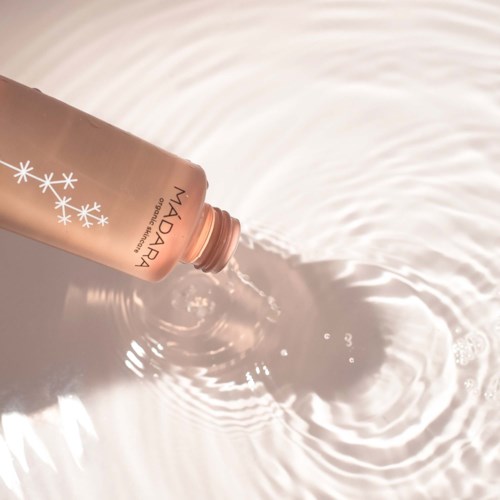 A pink bottle labeled 'Madara' pours liquid into water, creating ripples and bubbles on the surface.