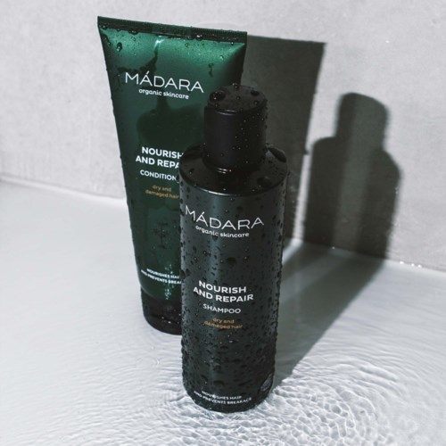 Two black bottles labeled 'MÁDARA' contain shampoo and conditioner, placed on a wet surface with droplets and a shadow behind.