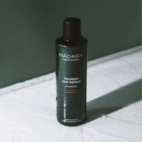 A black bottle of Madara nourishing shampoo with water droplets, placed on a white textured surface with a shadow.