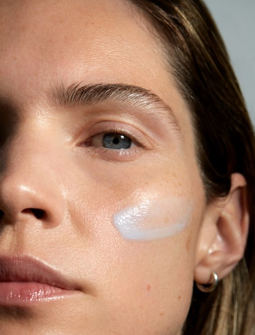 Close-up of a woman's face with a white skincare product on her cheek, blue eye, and earrings.