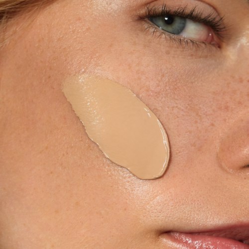 A woman with blue eyes has beige cream or foundation on her cheek, near her eye.