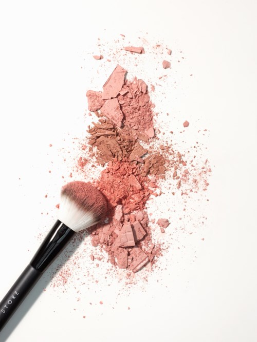 En makeupborste ligger på rosa puderrouge med utspridda bitar, på en vit bakgrund.
