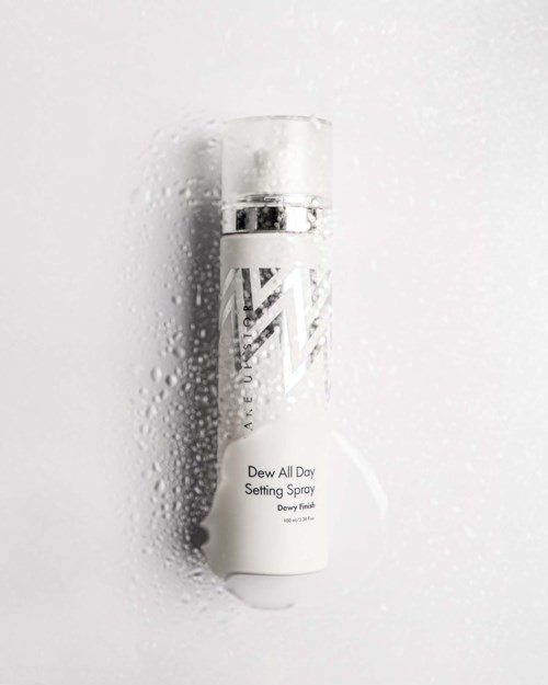 A white spray bottle with a silver cap, labeled Dewy All Day Setting Spray, surrounded by water droplets.