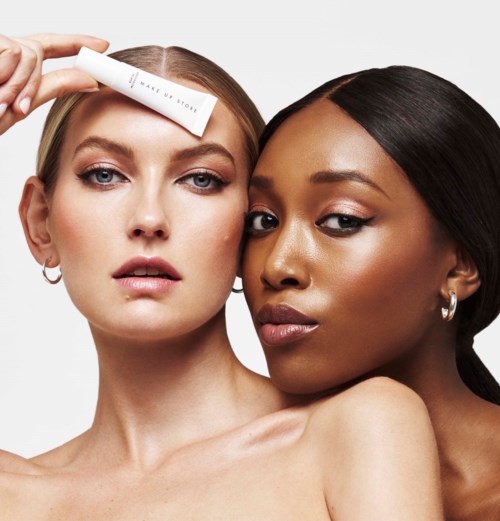 Zwei Frauen mit Make-up und Schmuck, eine hält ein Produkt auf ihrer Stirn, nah beieinander vor einfachem Hintergrund.