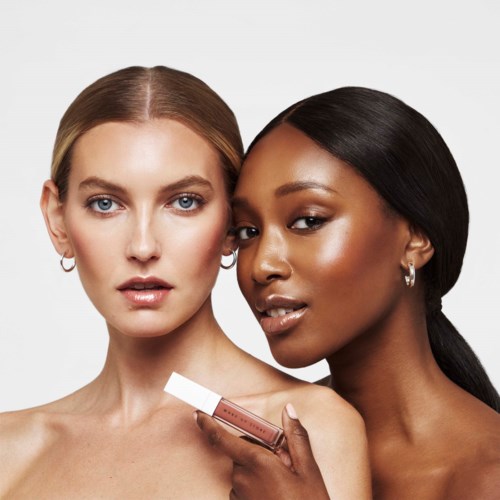 Twee vrouwen met verschillende huidschijnen poseren dicht bij elkaar, de ene houdt een lippenstift dicht bij de ander, op een witte achtergrond.