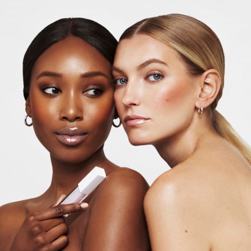 Twee vrouwen met verschillende huidtinten kijken naar de camera, de ene houdt een wit voorwerp, ze lijken zelfverzekerd en dichtbij.