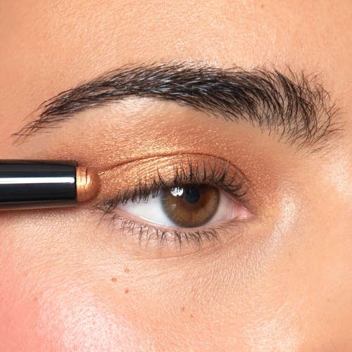 Een persoon brengt gouden oogschaduw aan met een kwast op het ooglid, dicht bij de wimpers. Het oog is bruin en de make-up is gedetailleerd.