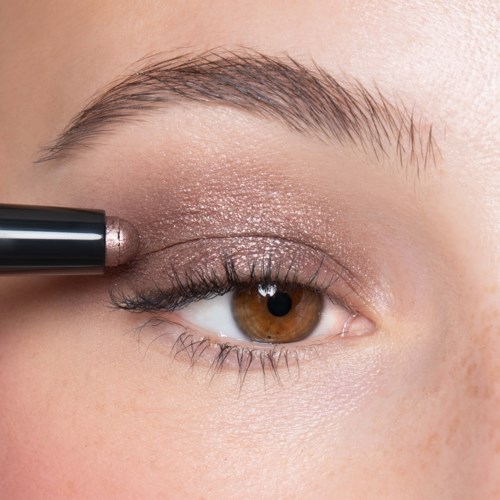 Eine Person trägt schimmernden Lidschatten mit einem Make-up-Stift auf. Sie haben braune Augen und helle Haut.