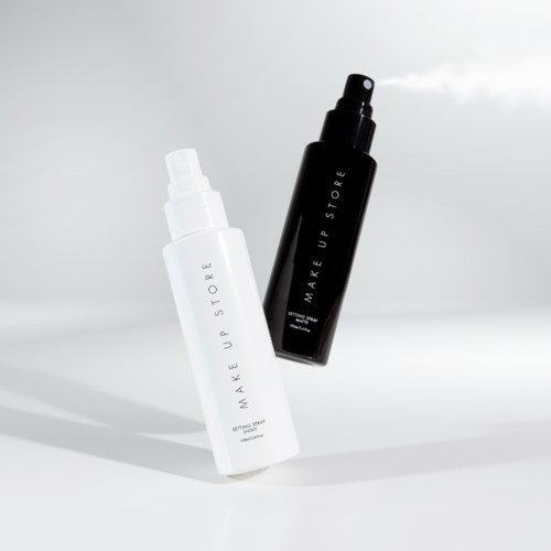 Två sprayflaskor, en vita och en svarta, märkta 'MAKE UP STORE,' står på en enkel bakgrund.