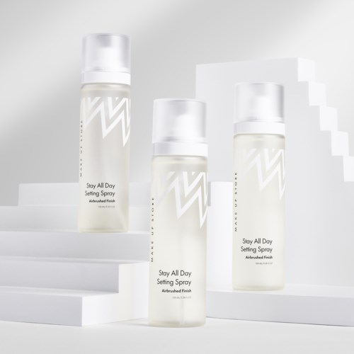 Drei Flaschen mit Setting Spray stehen auf weißen Stufen vor modernem Hintergrund. Sie sind mit "Stay All Day" beschriftet.