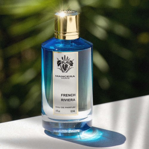 Blauwe parfumfles met een gouden dop, gelabeld 'French Riviera' en een leeuwenlogo, op een zonnige oppervlakte.
