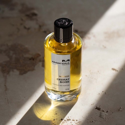 Doorzichtige glazen fles met gele vloeistof en zwart deksel, gemarkeerd "Cedar Boise". Staat op een stoffig oppervlak in zonlicht.
