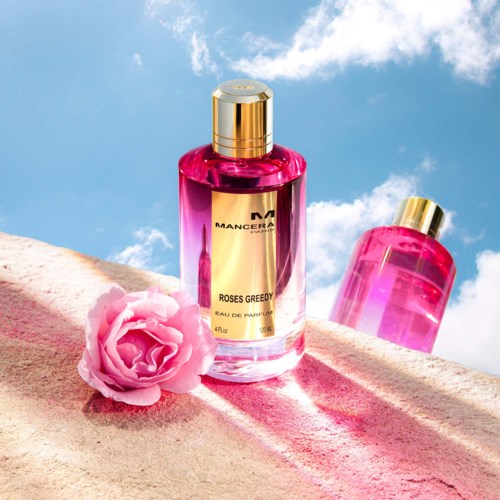 Twee roze parfumflesjes met een roze roos op zachte, roze stof onder een blauwe lucht. Eén fles is gemarkeerd 'MANCERA'.