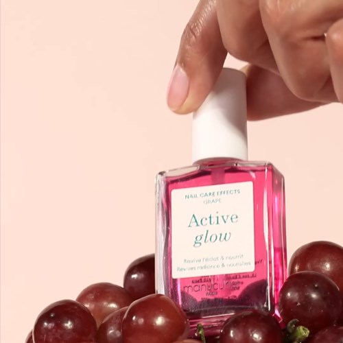A hand holds a pink nail polish bottle labeled 'Active glow' among grapes.