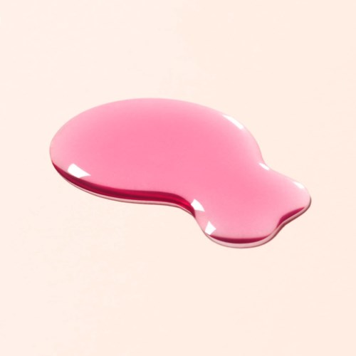 A pink liquid drop spread out on a light surface with shiny highlights.