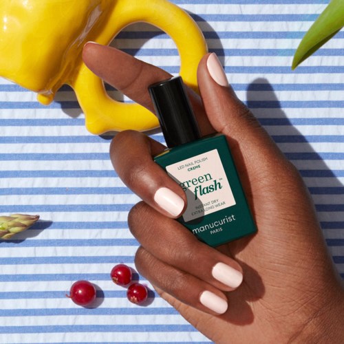 A hand holds a green nail polish bottle with white nails, surrounded by yellow bell pepper, red berries, and a leaf.