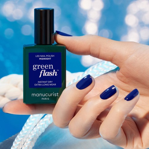 Green nail polish bottle with dark blue nails and a hand holding it, background of shiny blue lights.