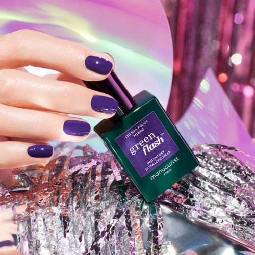 A person's hand with purple nails holds a green nail polish bottle labeled "Green Flash" on shiny silver and pink decorations.