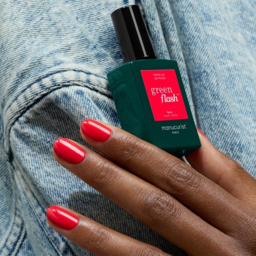 A person holds a green bottle with bright red nails, standing near blue denim fabric. The bottle is labeled Green Flash.