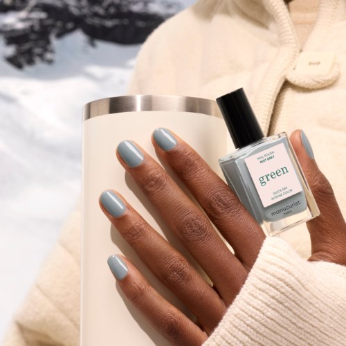 A person with dark skin shows nails painted light blue, holding a white tumbler and a small green nail polish bottle.