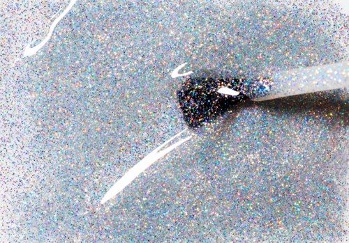 A sparkly, colorful surface with a white stick and small white lines, likely a close-up of nail polish application.