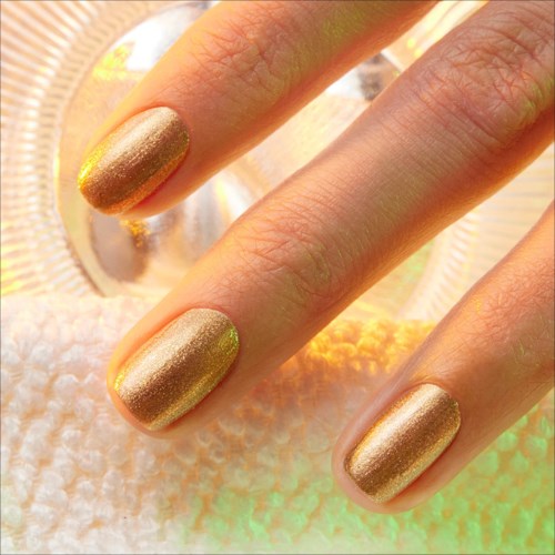 Three fingers with shiny, gold-colored nails resting on a soft, textured surface.