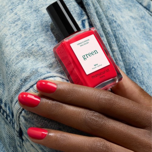 A hand with bright pink nails holding a red nail polish bottle labeled green. The person wears denim clothes.