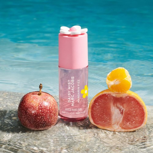 Maracuja fragrance mist with pink cap, next to an apple and half a grapefruit, on a sandy surface by water.