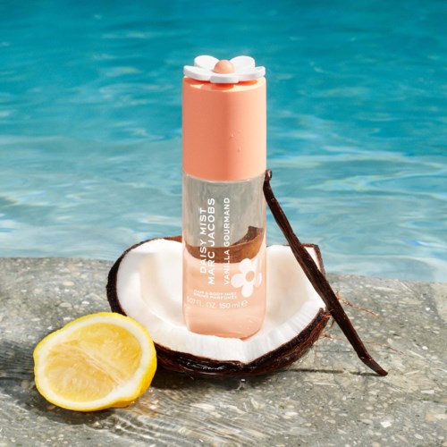 A pink lip balm tube with a white flower design, placed on a coconut shell with a lemon slice nearby, by water.