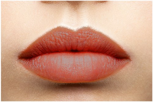Close-up of full lips with peach-colored lipstick and smooth skin. Bright lighting highlights lip texture.