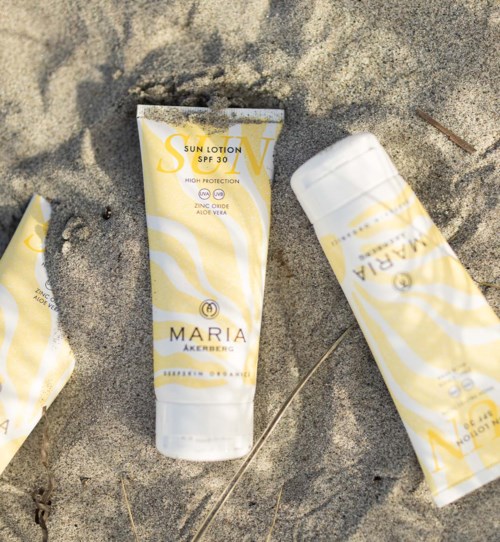 Sun lotion tubes are lying on sandy ground, some partially buried. They are yellow and white with brand and SPF label.