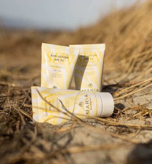 Three tubes of sun lotion on dry grass with a blurred background. The tubes are yellow and white, labeled 'Sun Lotion SPF 30' and 'Maria'.