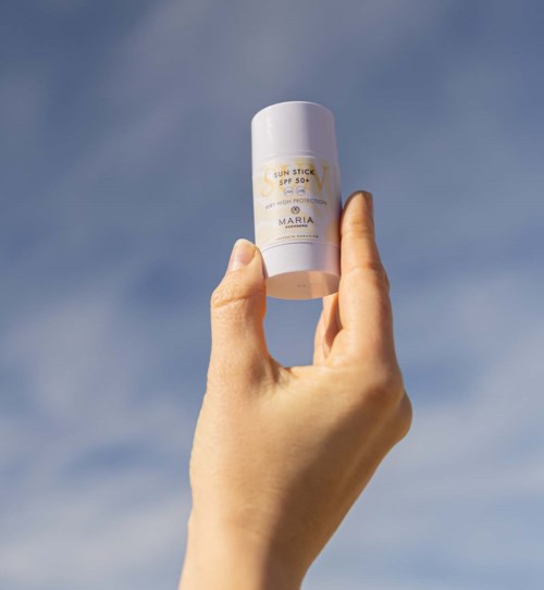 Een hand houdt een witte zonnebrandstick voor een blauwe hemel.