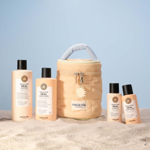 Vier Flaschen Haarpflegeprodukte und eine beige Tasche mit Griff stehen auf einer sandigen Oberfläche vor einem hellblauen Hintergrund.