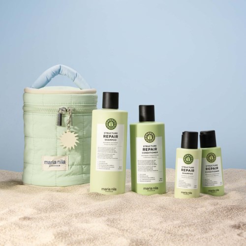 Vier Flaschen Haarpflegeprodukte und eine mintgrüne Reisetasche auf Sand vor hellem blauen Hintergrund.