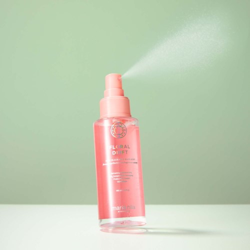 Pink spray bottle releasing mist on a green background. It is labeled Floral Drift by Marini Nila.