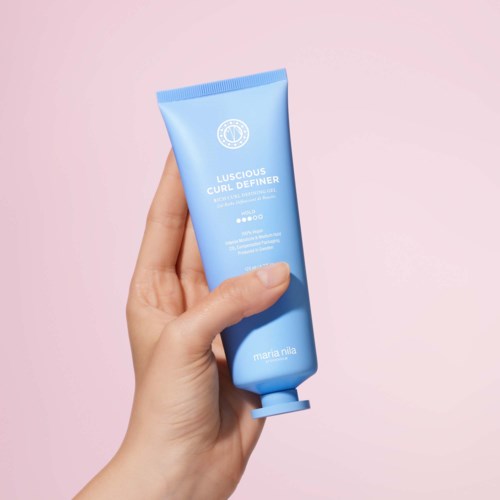 A hand holds a blue tube labeled Luscious Cure Definer by Maria Mila against a pink background.