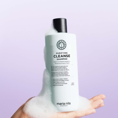 A white shampoo bottle with black cap and bubbles on it, held in a hand against a light background.