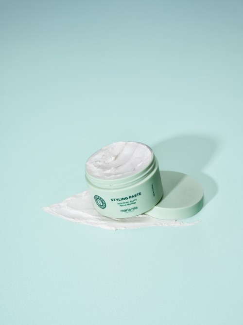 Ein runder mintgrüner Behälter mit Stylingpaste und etwas Produkt verschüttet auf hellblauem Hintergrund.