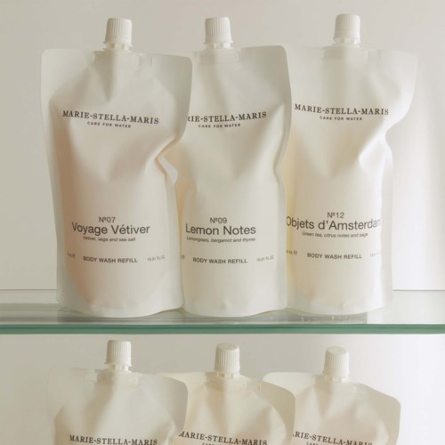 Sex vita påfyllningspåsar med kroppstvål, märkta med olika dofter och nummer, på ett glashylla.