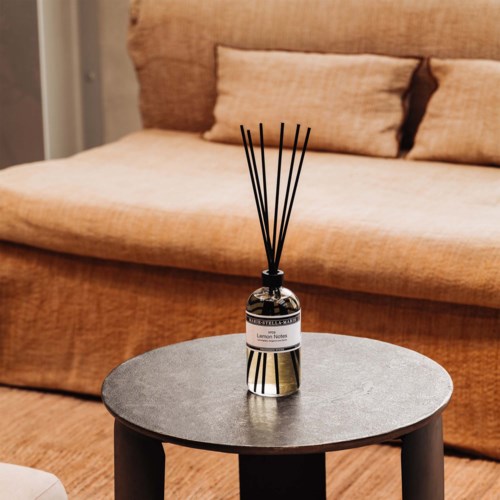 Een diffuser met zwarte stokjes op een klein ronde grijze tafel in een gezellige woonkamer.