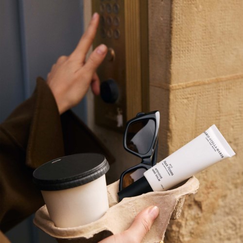 Person trycker på porttelefonen, håller kaffe, solglasögon och hudvårdsprodukter i ett papper.