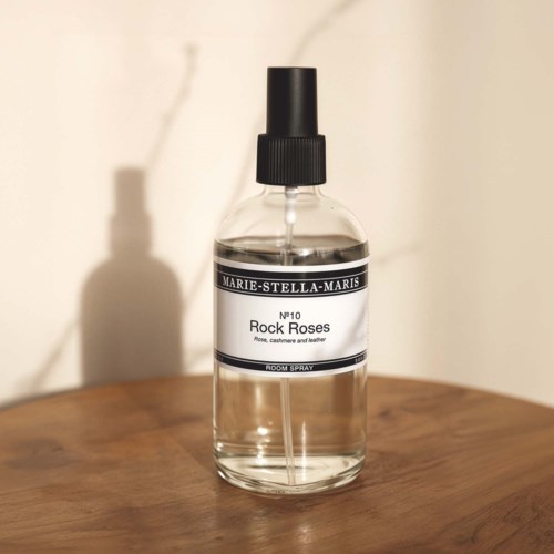A clear room spray bottle labeled Rock Roses, with a spray nozzle, on a wooden surface.