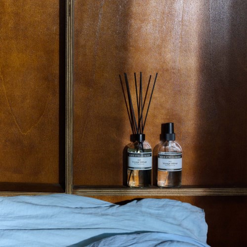 Twee glazen flessen met zwarte deksels en een reed diffuser staan op een houten oppervlak tegen een houten muur.