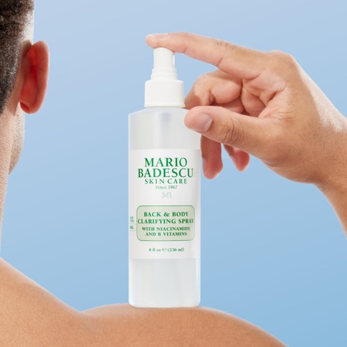 Käsi suihkuttaa Mario Badescu -ihonhoitosuihkeen henkilön olkapäälle. Pullo on valkoinen vihreällä tekstillä.