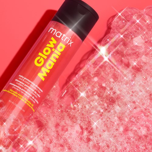 Eine rote Flasche mit der Aufschrift 'Glow Mania' in Gelb und Weiß, neben einer funkelnden, rosa, flauschigen Oberfläche.